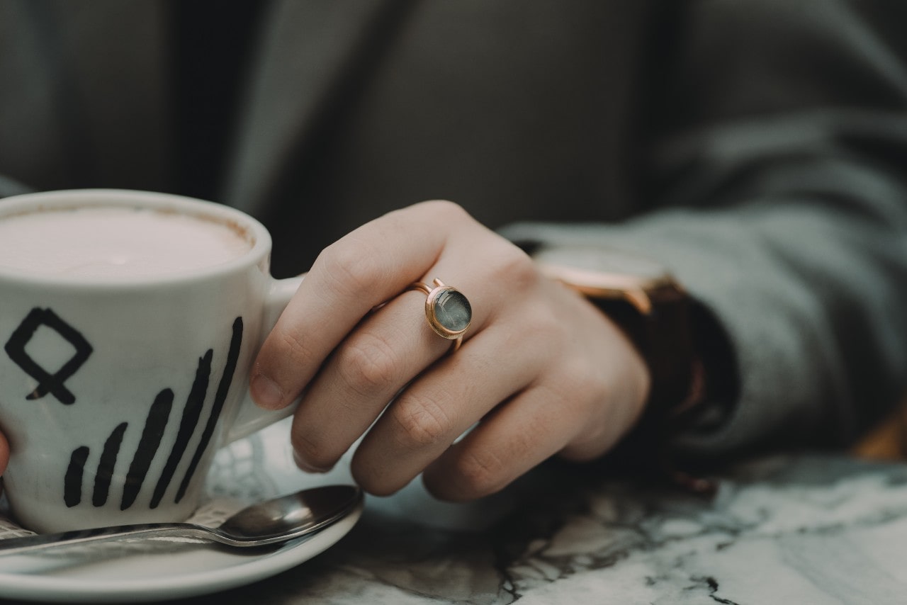 Bezel-set gemstone ring sits on woman’s middle finger as she drinks coffee