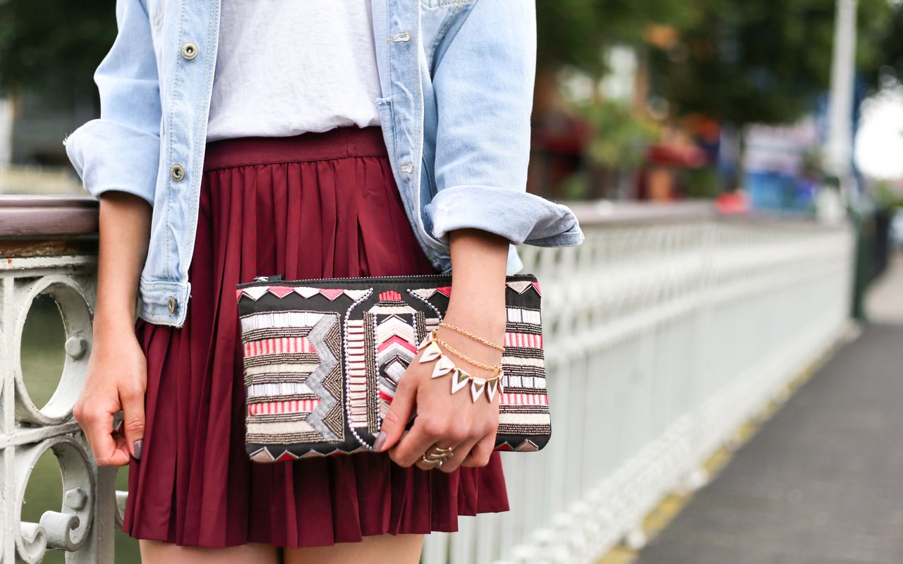 Standing by a decorative wall, a woman is waiting for her date in a maroon mini skirt, denim overshirt, and a graphic clutch that matches her bracelets and fashion rings