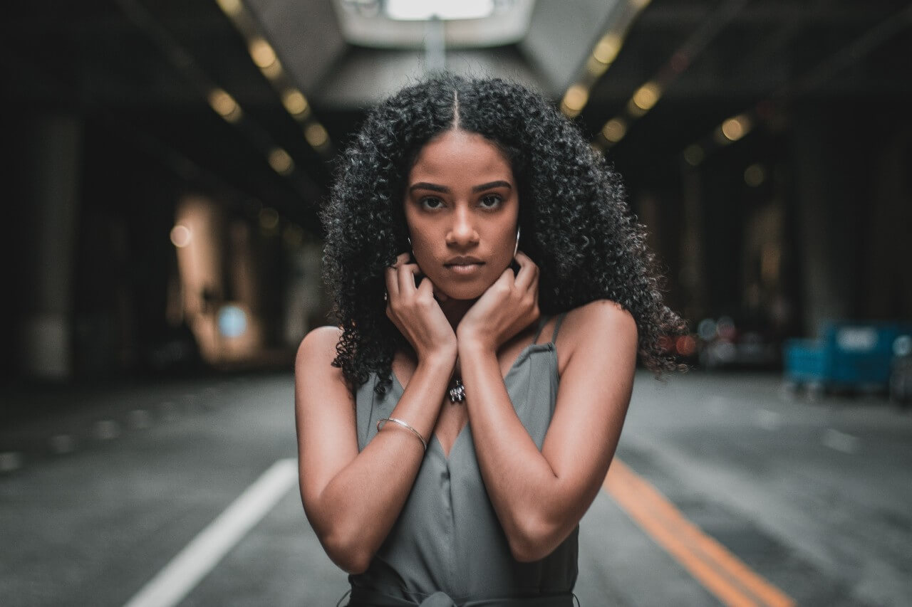 Standing in a city street, a woman stares into the camera while wearing a gray romper, single silver bangle on her wrist, large hoop earrings, and a statement pendant