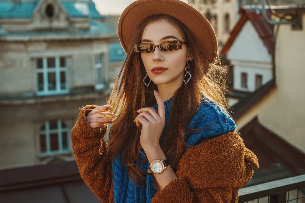 Woman standing on a balcony wearing a brown hat that matches their coat along with sunglasses, drop geometric earrings, and a watch