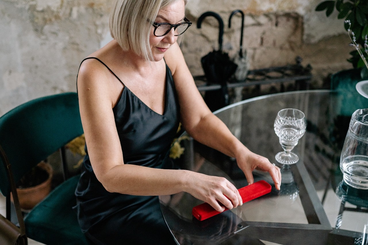 Woman in a black dress about to open a long red jewelry box while seated at the dining room table at a restaurant