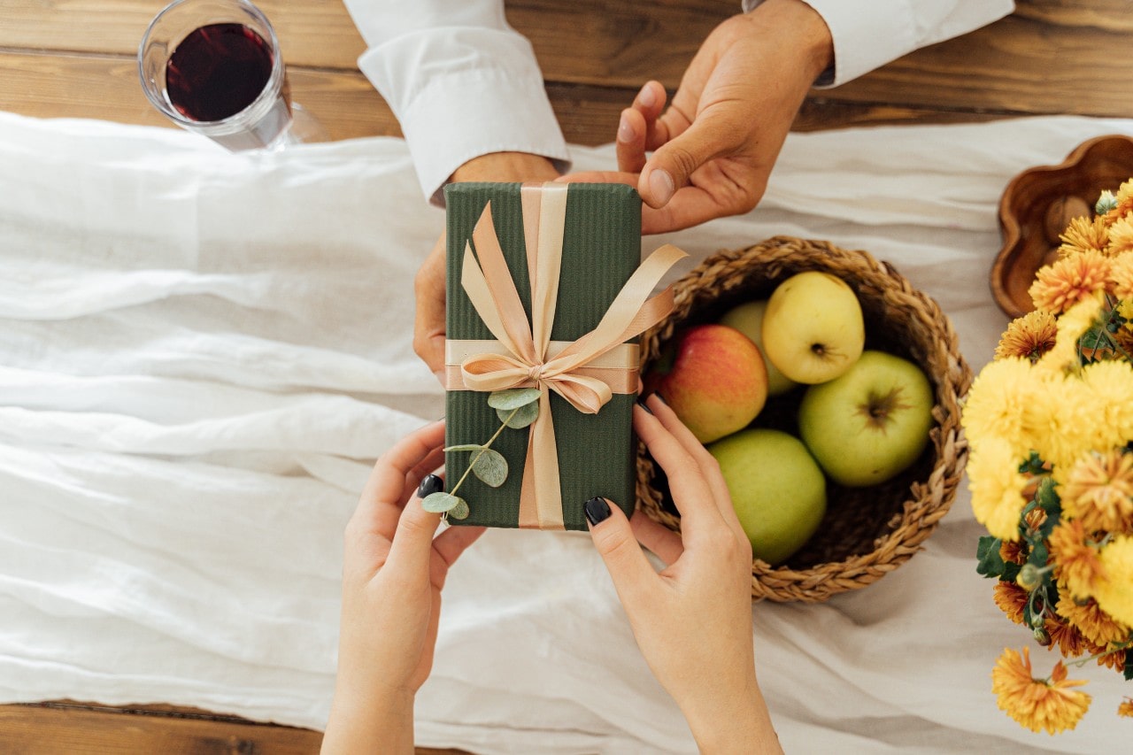 A man and woman exchanging an olive-colored package with a pale pink bow over a table with a white table runner, a basket of apples, and a pot of flowers