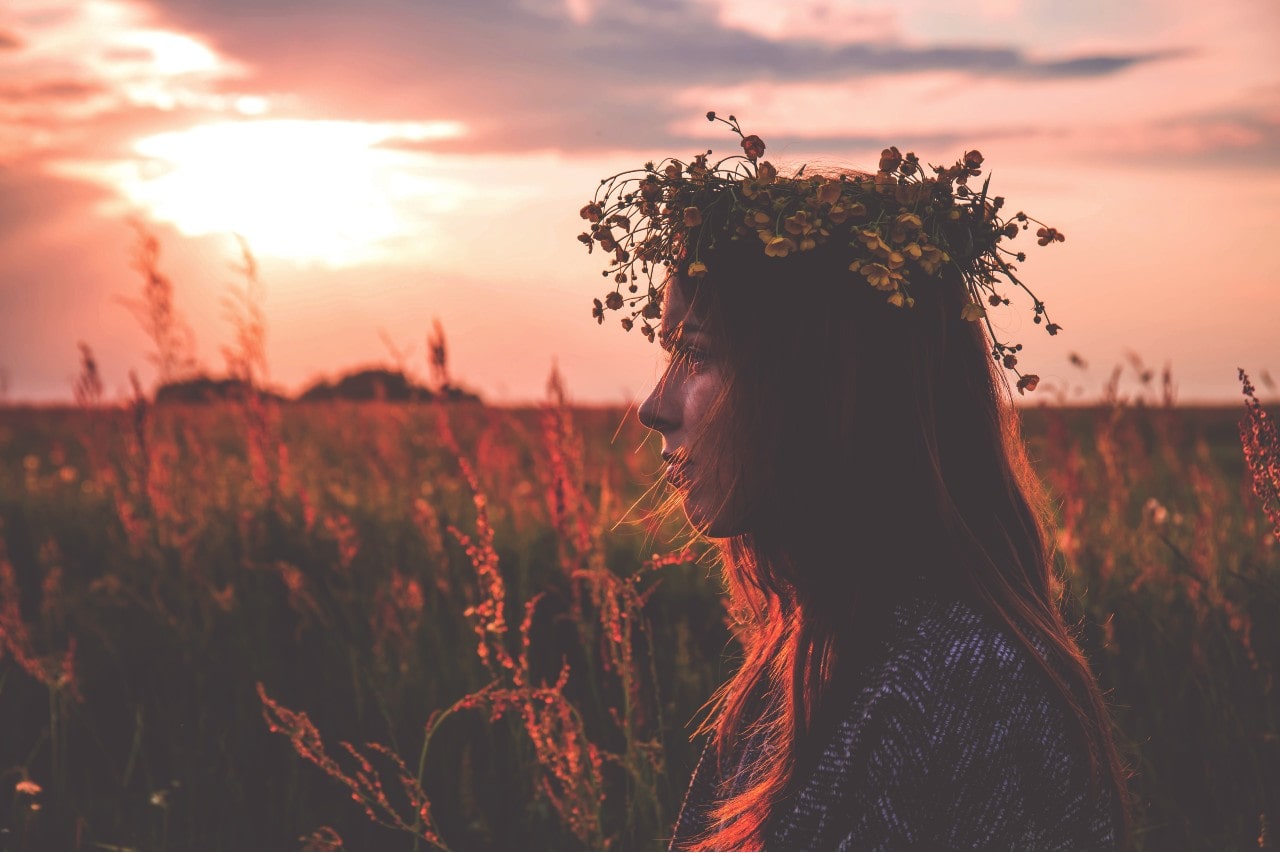 A woman sitting outside in the early morning light, surround by fields and flowers, wearing a flower crown