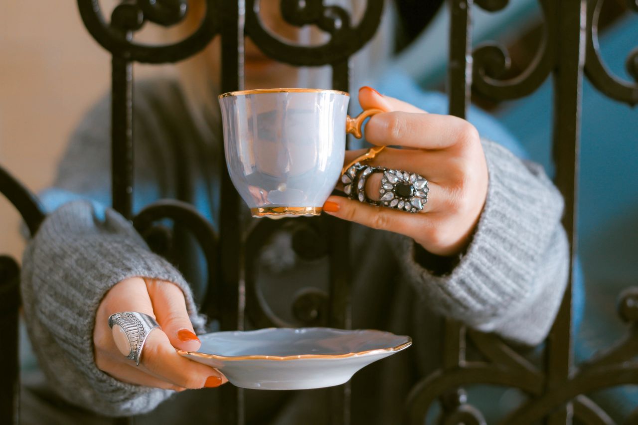 A woman holding a teacup and saucer through an iron gate wearing bold gemstone fashion rings, one has a floral design on it
