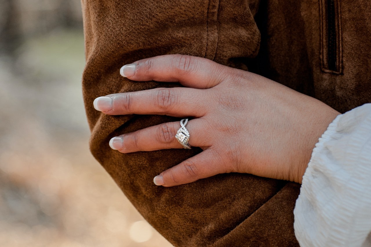 A diamond and moissanite ring on a woman’s hand resting on the elbow of her partner wearing a tan suede jacket