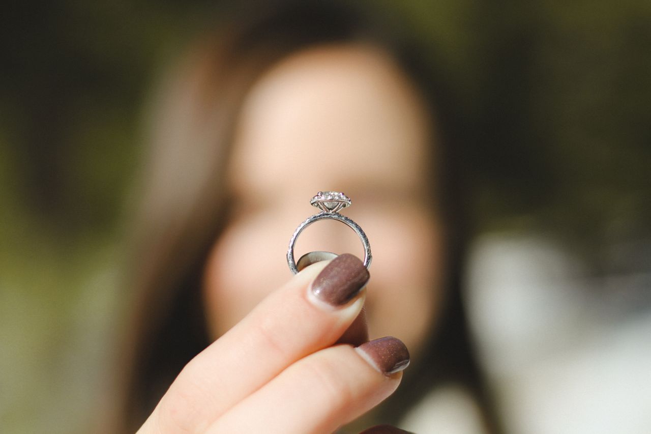A close up of a side view of a halo engagement ring that a woman is holding between her fingers