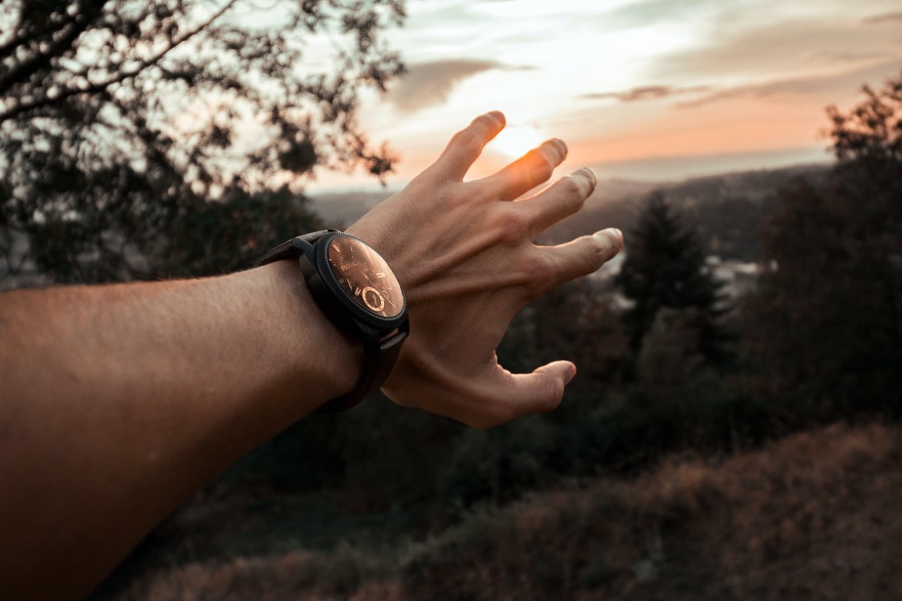 A man’s outstretched hand reaching towards a sunny horizon and wearing a black sports watch