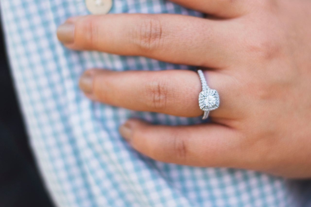 A woman wears a side stone engagement ring featuring a halo and a round cut diamond