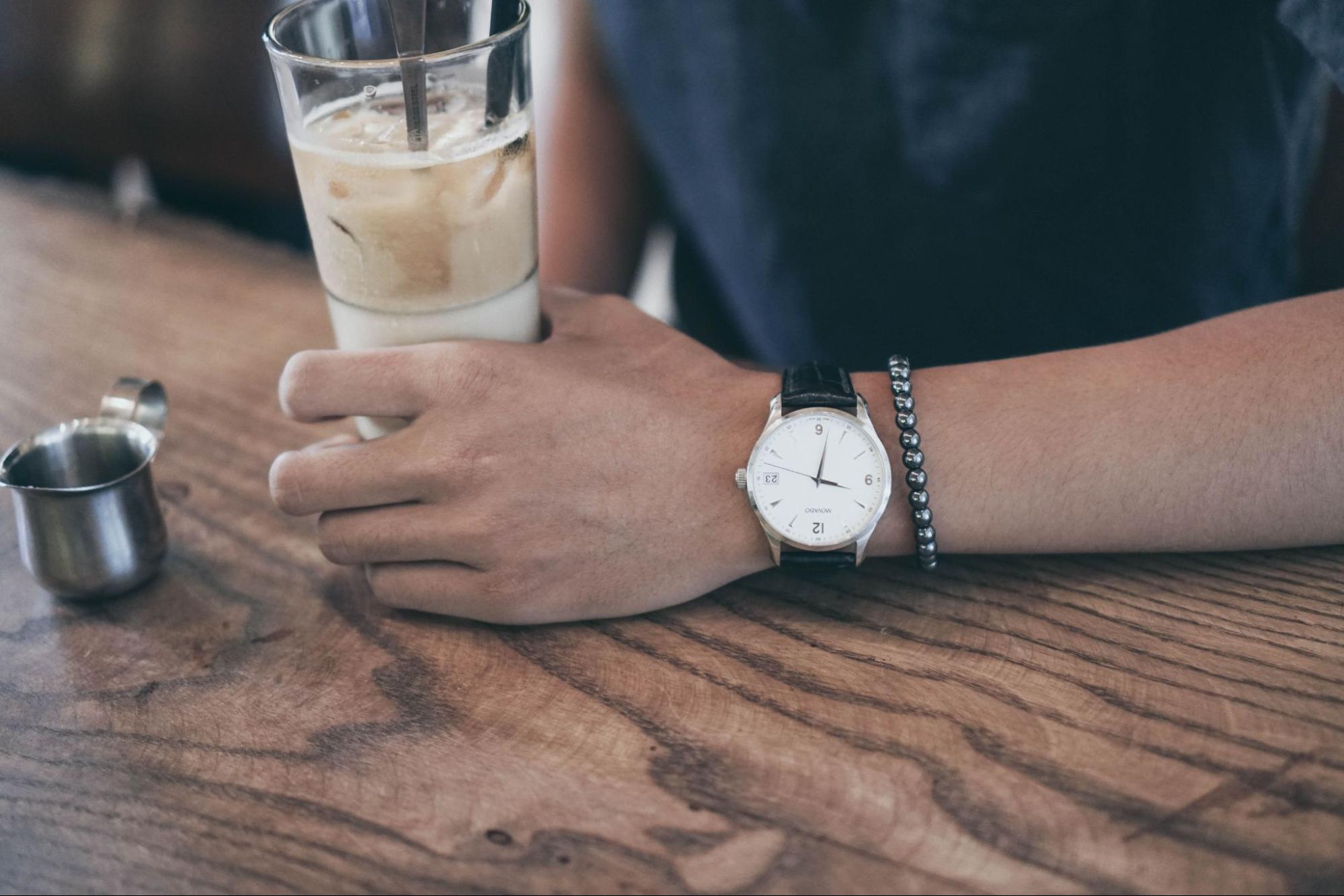 Close up image of a man holding an iced coffee, wearing a silver and white watch and a black beaded bracelet