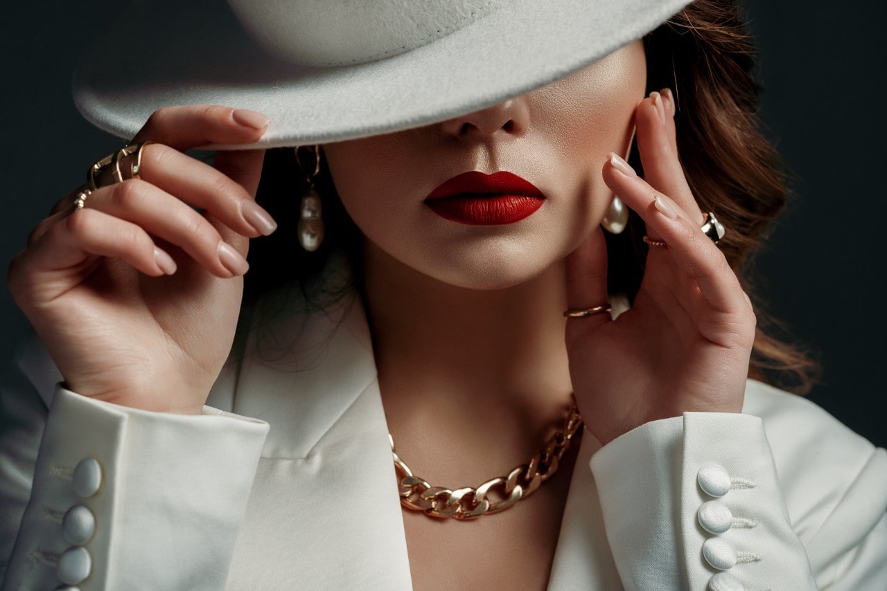 A woman wearing designer jewelry covers her eyes with her white fedora