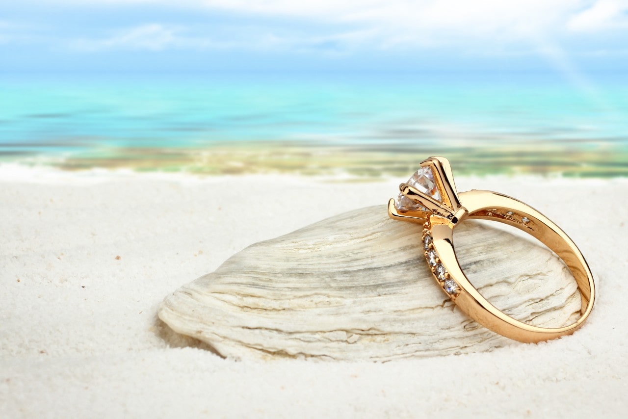 A gold sidestone ring sits on a seashell on the beach.