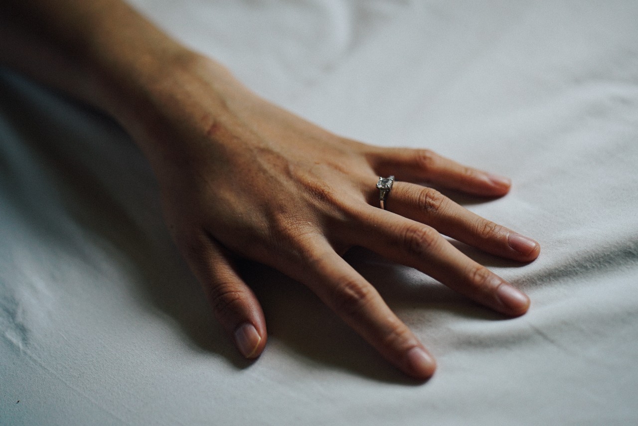 A closeup of a hand with a three-stone engagement ring stretches on a bed.