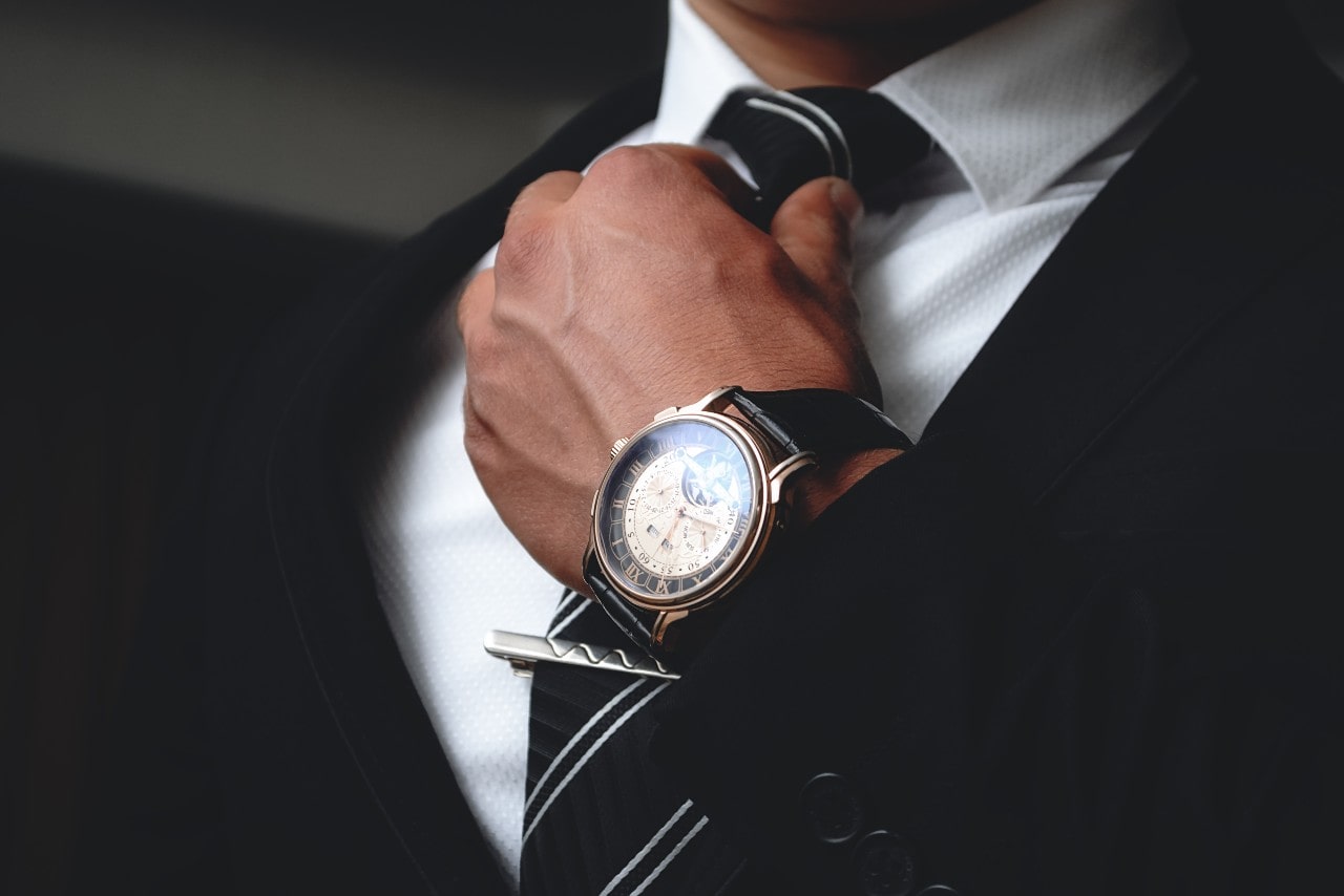 Close-up of a man in a black suit fixing his tie, showcasing black leather watch with gold plated bezel.
