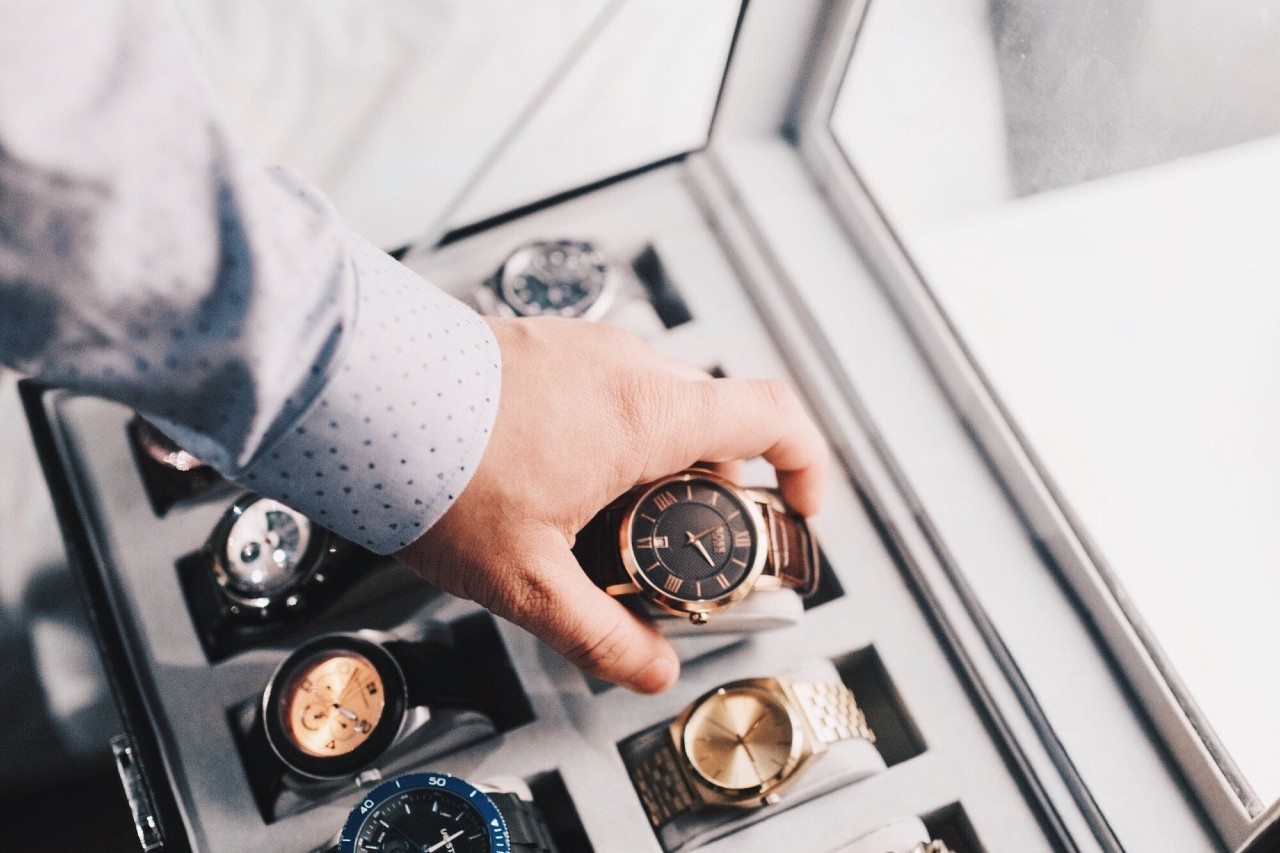A hand in a polka dot shirt selects a stylish brown and gold wristwatch from a display case filled with diverse, elegant watches by a window.