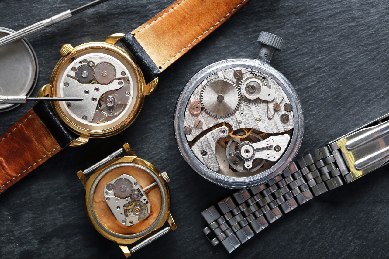 Three disassembled watch movements with gears and springs are laid out on a slate surface, flanked by watchmaker tools and a metal bracelet.
