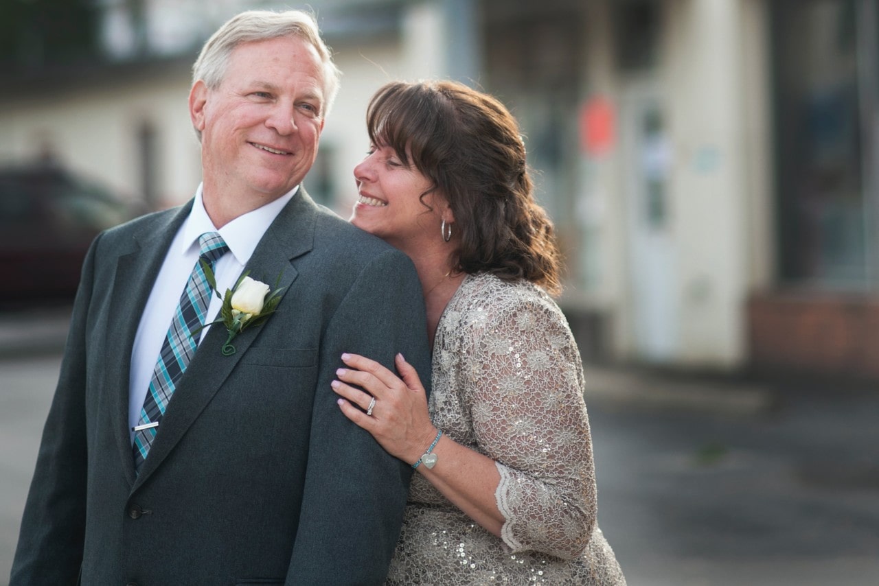 A smiling woman in a lace dress affectionately leans on an elderly man in a suit with a white rose boutonniere.