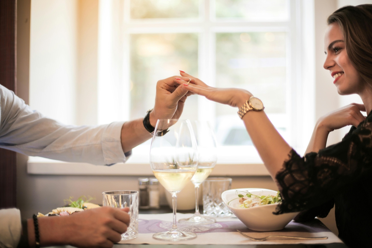 A man slides a ring on a woman's finger across a table set with wine glasses and a salad bowl, smiling warmly in a softly lit, romantic setting by a window.