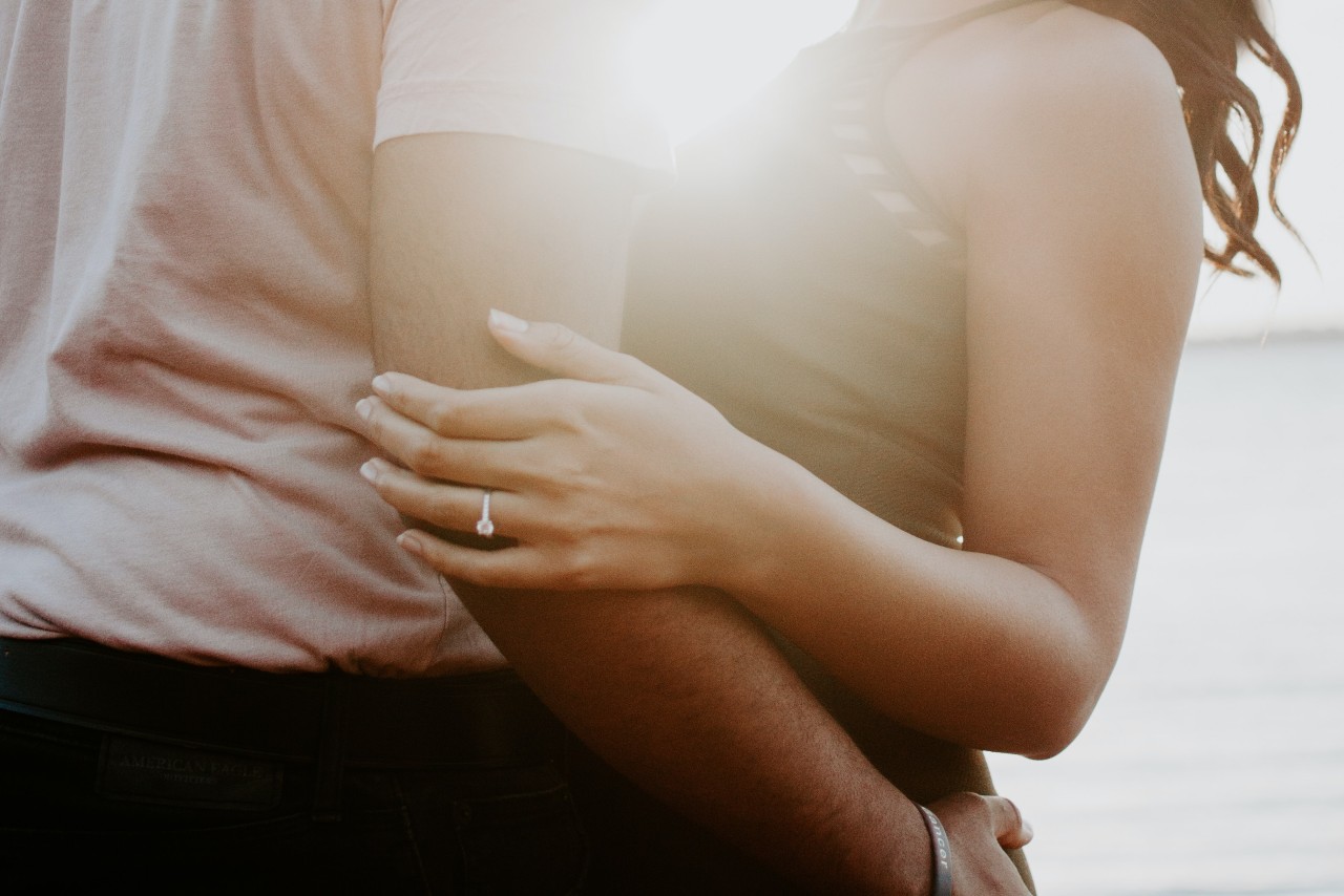 A close up of a couple embracing against a sunlight, showcasing woman&rsquo;s diamond engagement ring