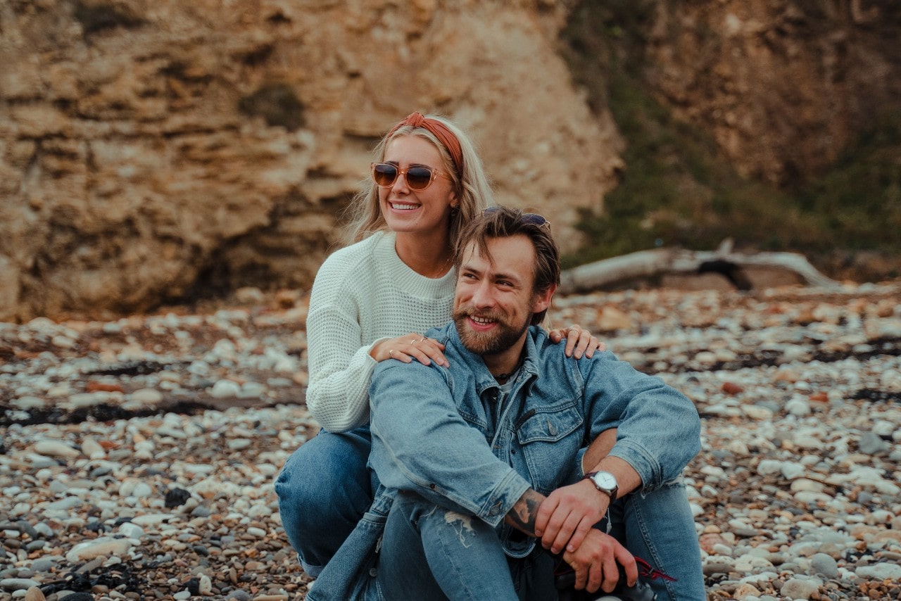 A smiling couple sits together on a rocky beach, with the woman resting her hands on the man&rsquo;s shoulders as they enjoy the coastal scenery.