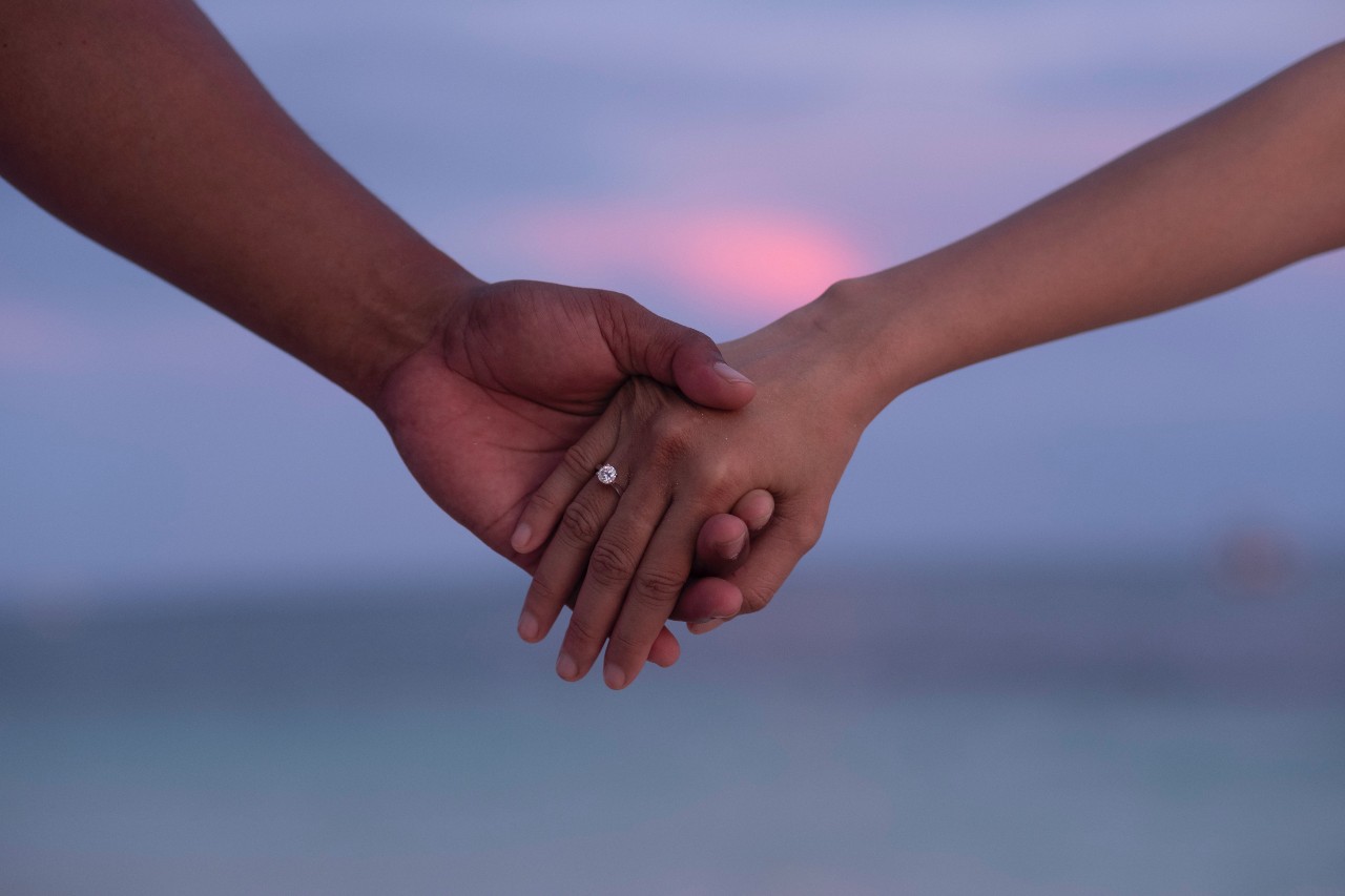 Two hands holding against a purple sunset, one hand adorned with solitaire diamond engagement ring