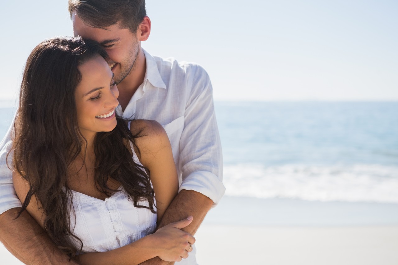 Couple in a white shirt and dress embracing in front of the ocean coast.