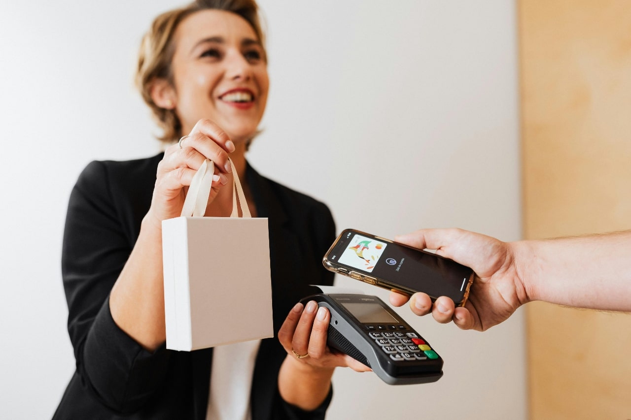 A smiling shop assistant hands over a small white shopping bag while a customer pays with a smartphone using contactless payment.