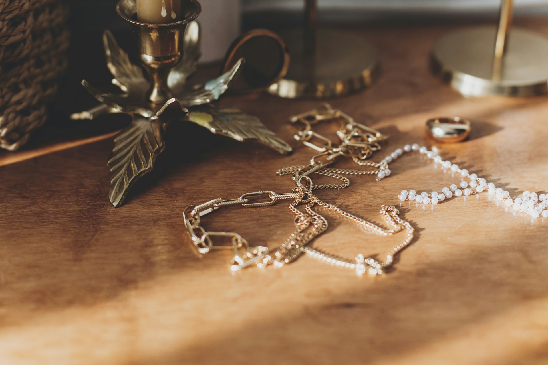 A collection of gold chains and a pearl necklace elegantly arranged on a wooden surface beside brass candle holders.