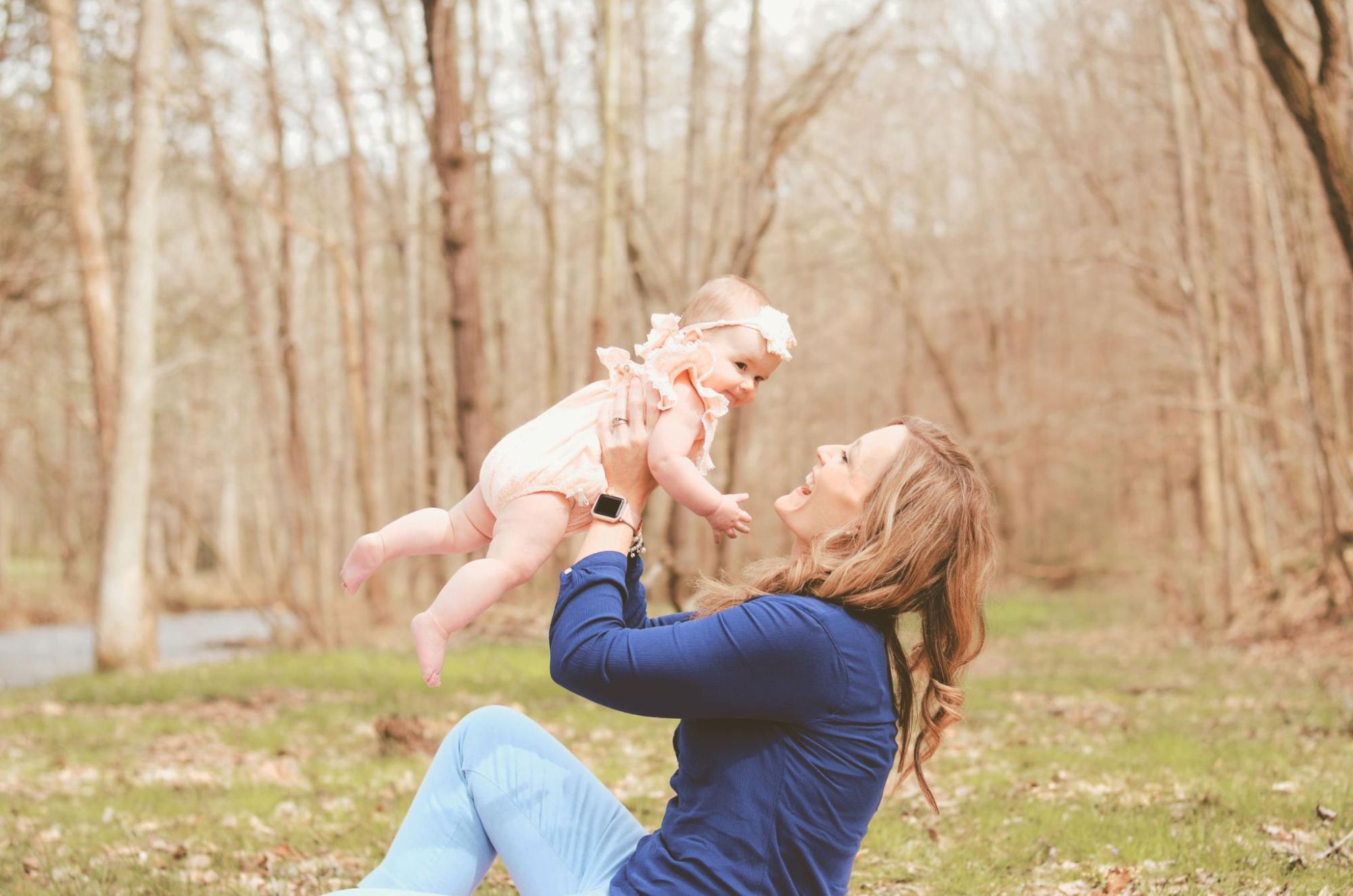 A woman in a blue shirt joyfully lifts a baby dressed in pink in an autumn forest.