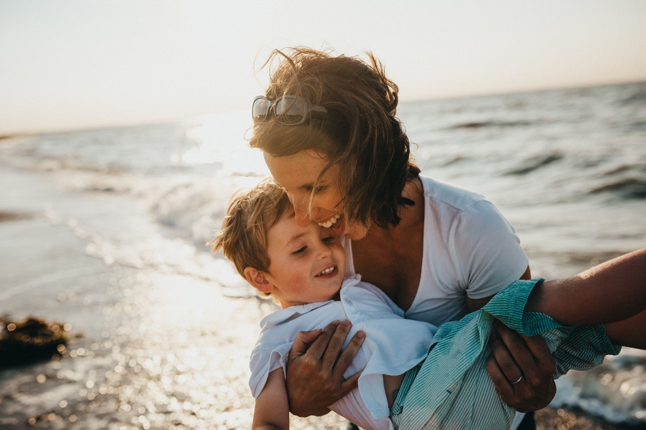 A joyful mother in a white top holds a smiling child by the ocean at sunset.