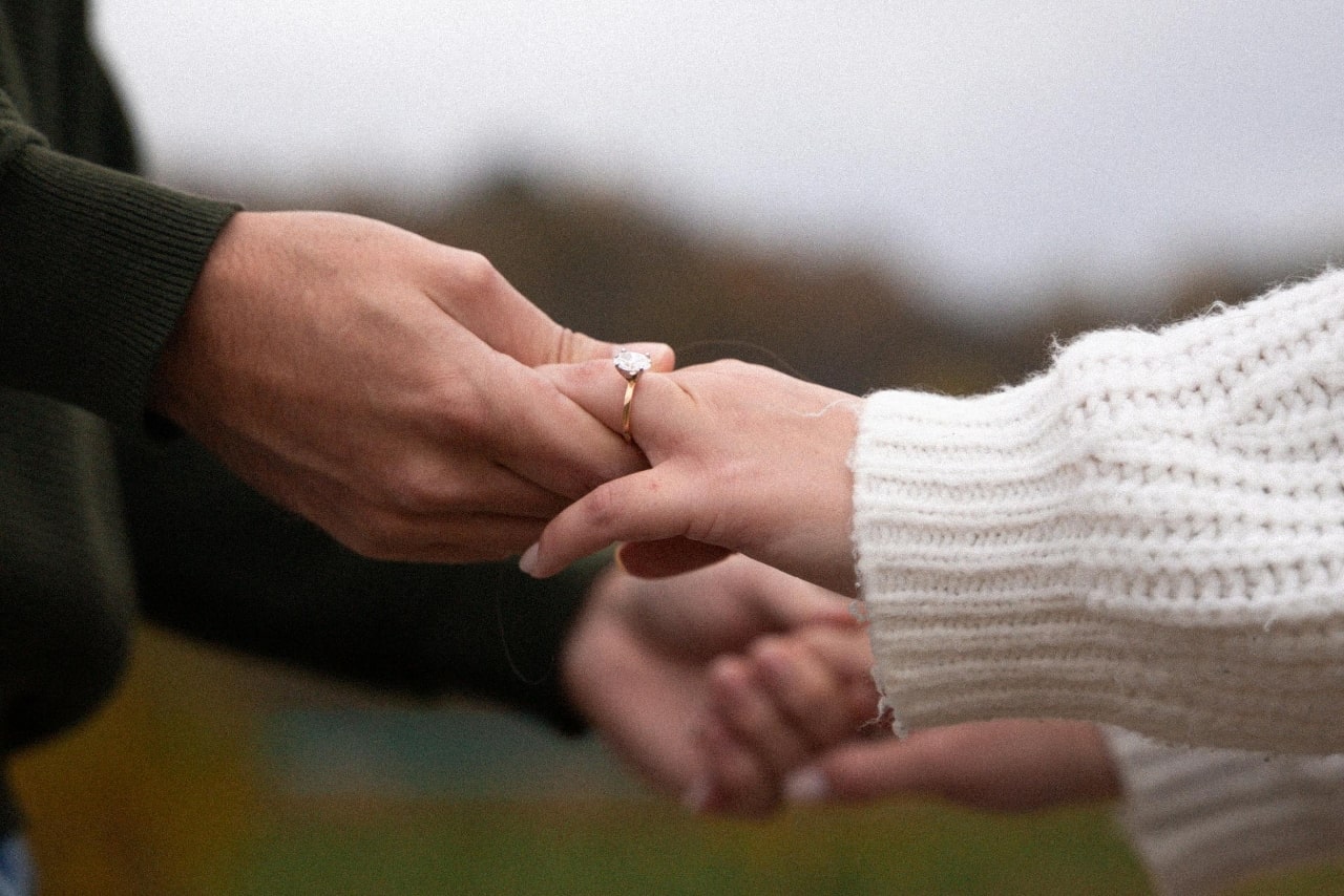 A close up of two hands, showcasing a yellow gold round cut diamond engagement ring.