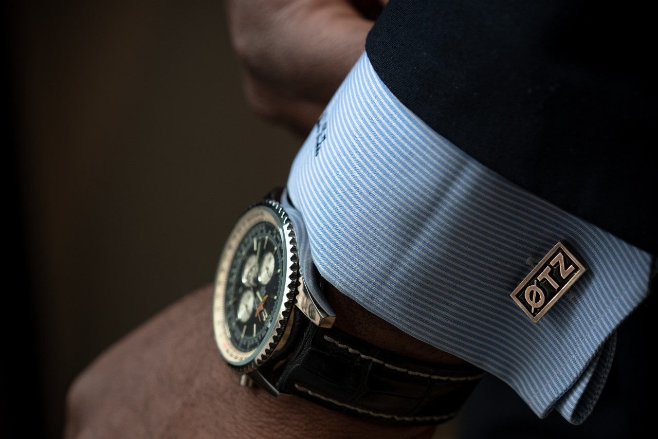A close-up of a man's wrist wearing a luxury chronograph watch with a black leather strap and a blue shirt with custom cufflinks.