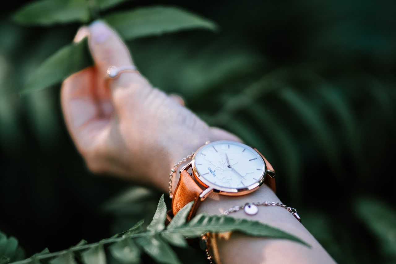 A woman's wrist adorned with a white-dial wristwatch, a delicate bracelet, and a ring, set against a lush green background of leaves.