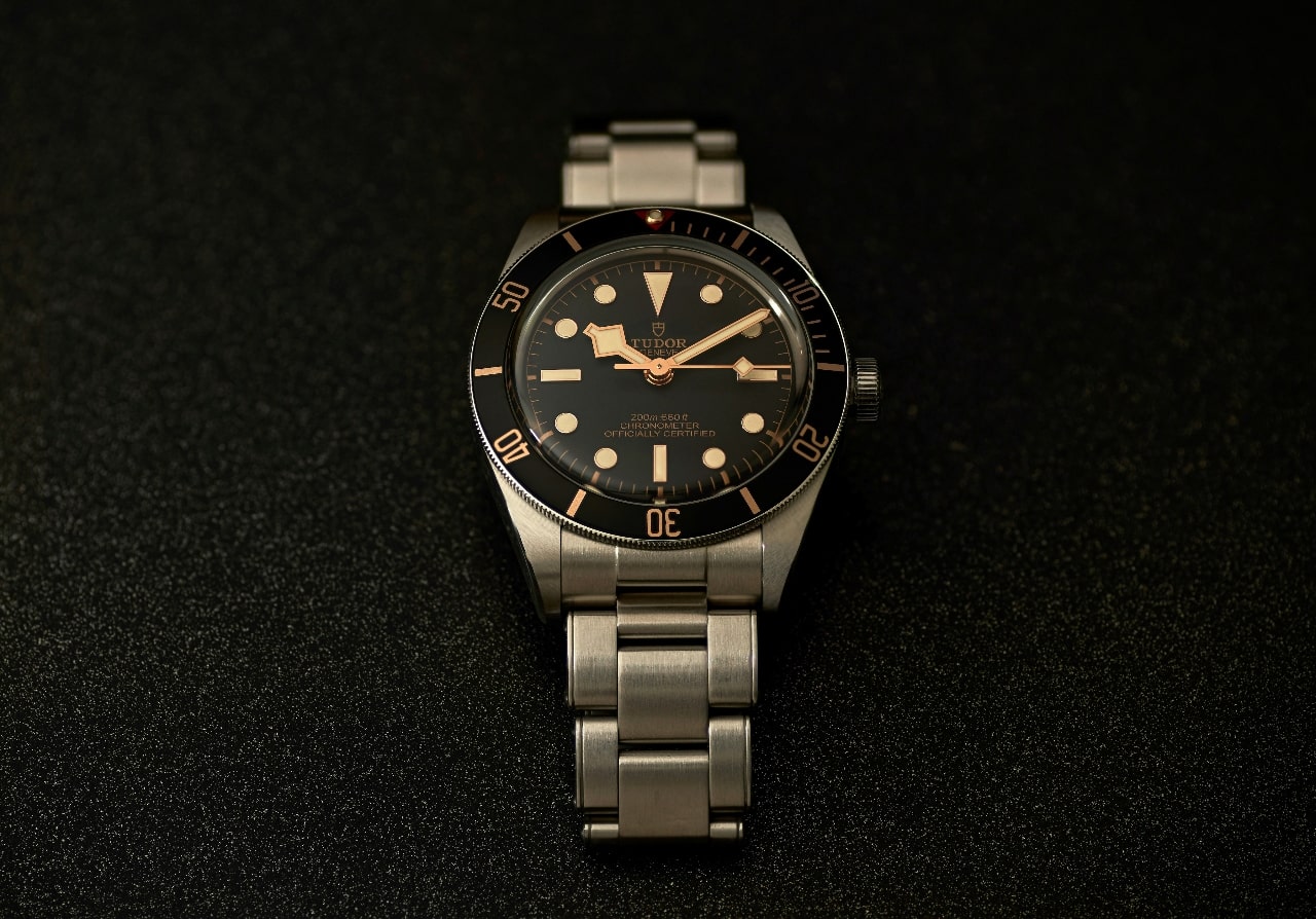 An angled top-down view of an elegant TUDOR watch displayed on a textured black backdrop.
