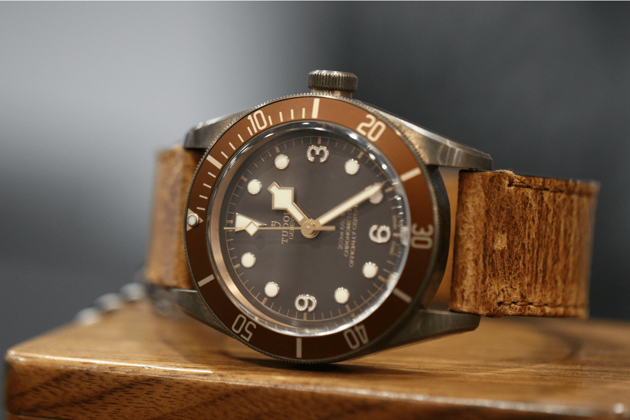 A close-up of a vintage-inspired TUDOR watch with a leather band displayed on its side on a wooden surface.