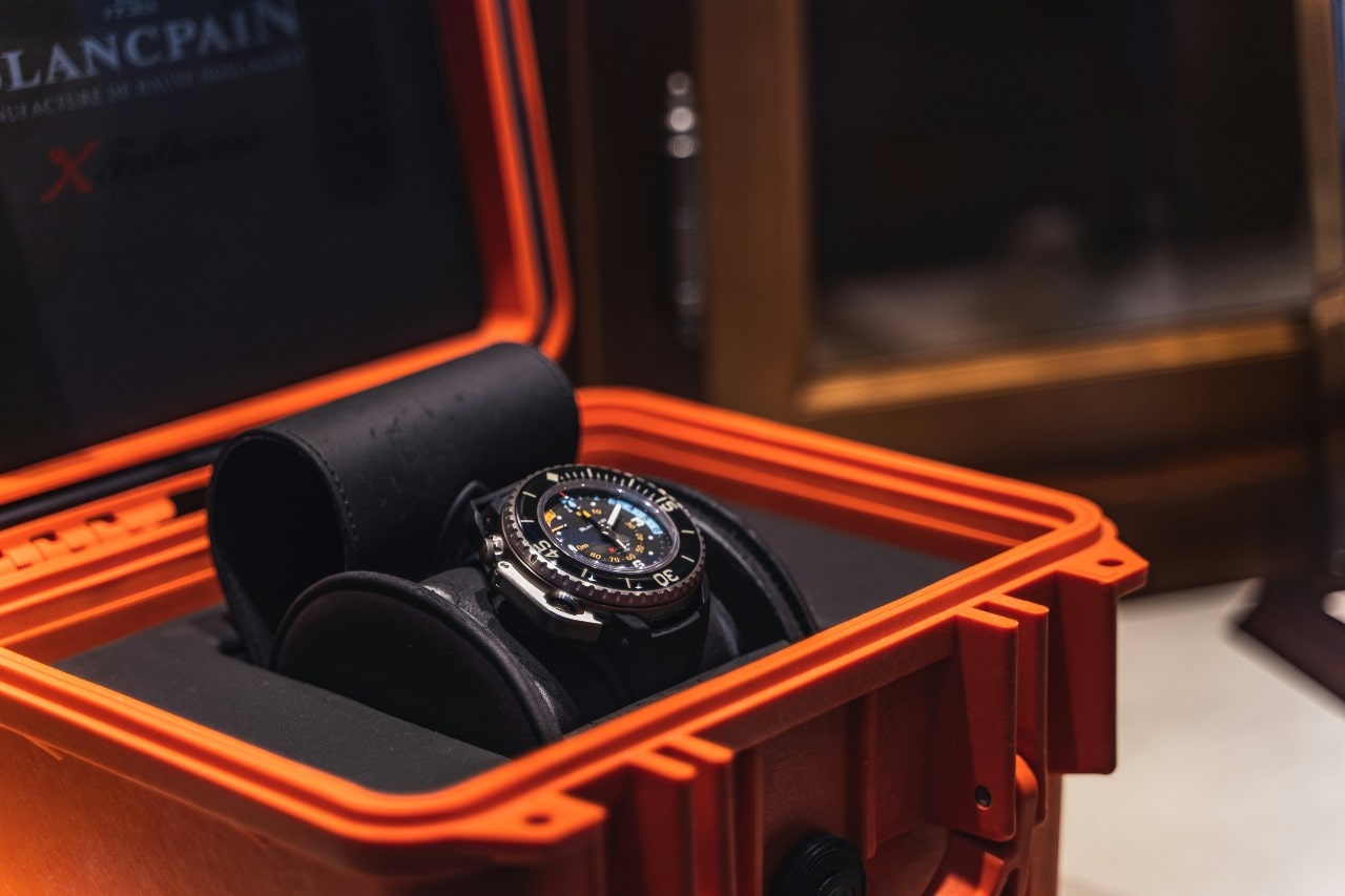 A black luxury watch is displayed in an open orange case.