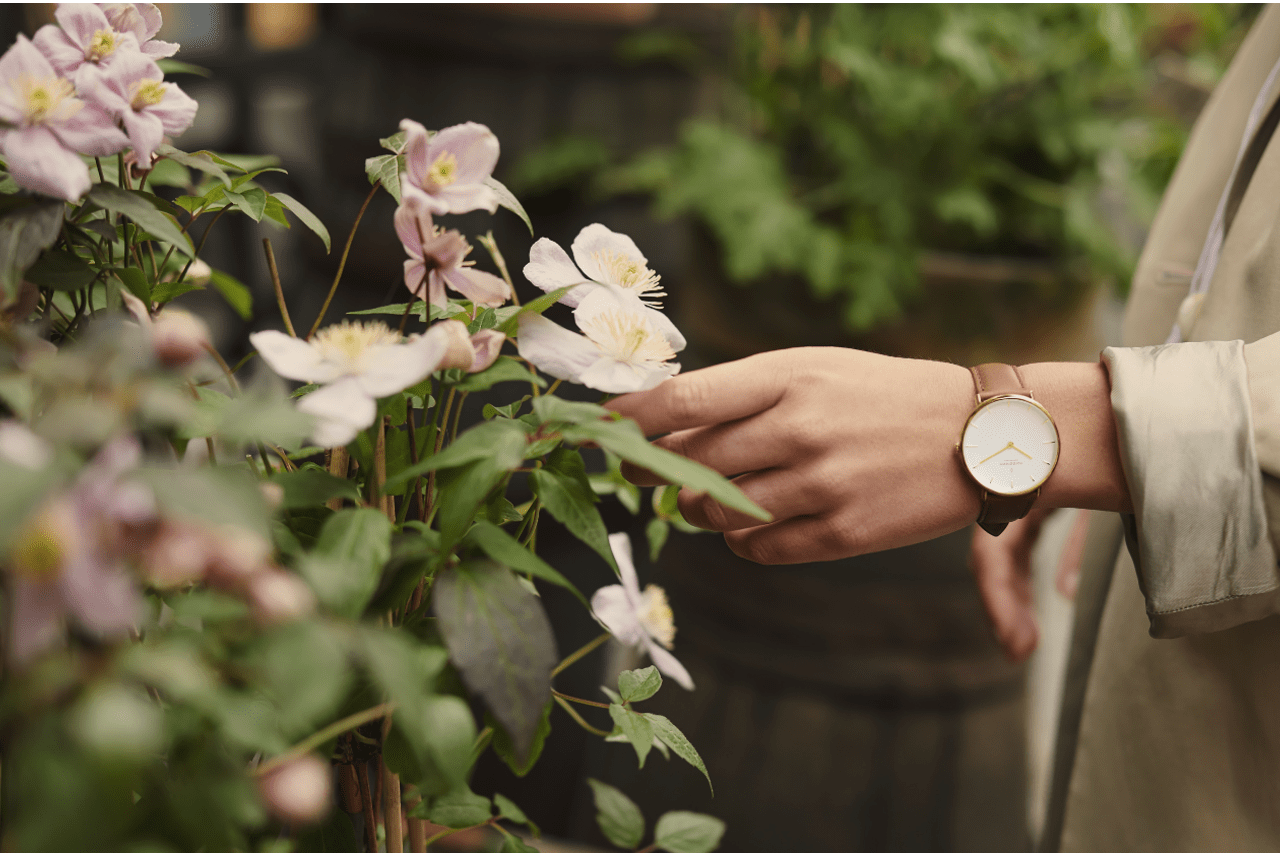 A hand wearing a minimalist watch touches delicate white flowers amidst lush greenery.