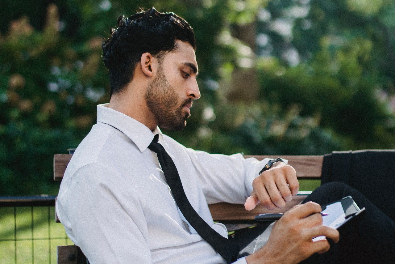 A man in white shirt and black tie sitting on a park bench checking his wristwatch against a blurred green background.
