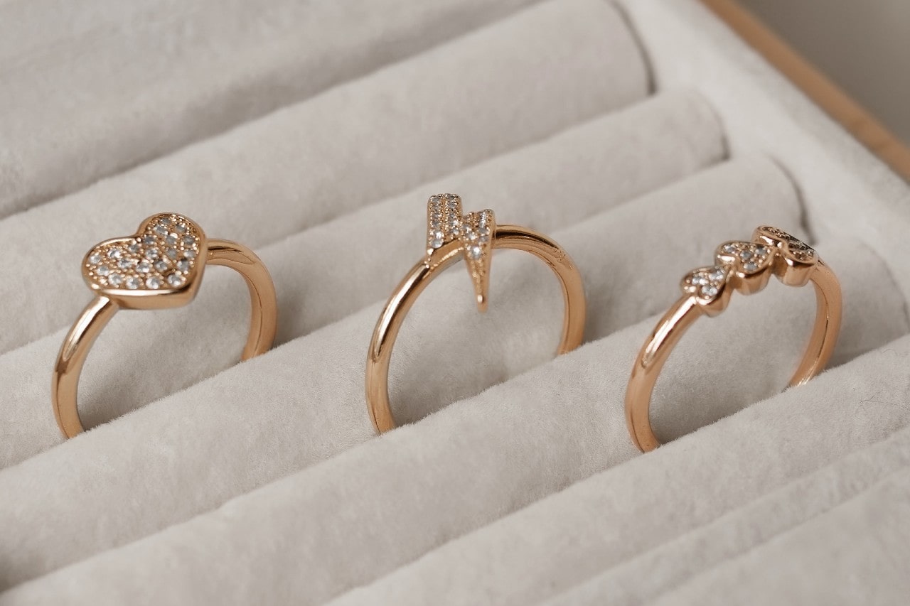 A close up of 3 yellow gold fashion rings with sparkling diamonds in a different shape – a heart, a lightning bolt, and a trio of round stones – rest in individual slots within a light-colored jewelry display.