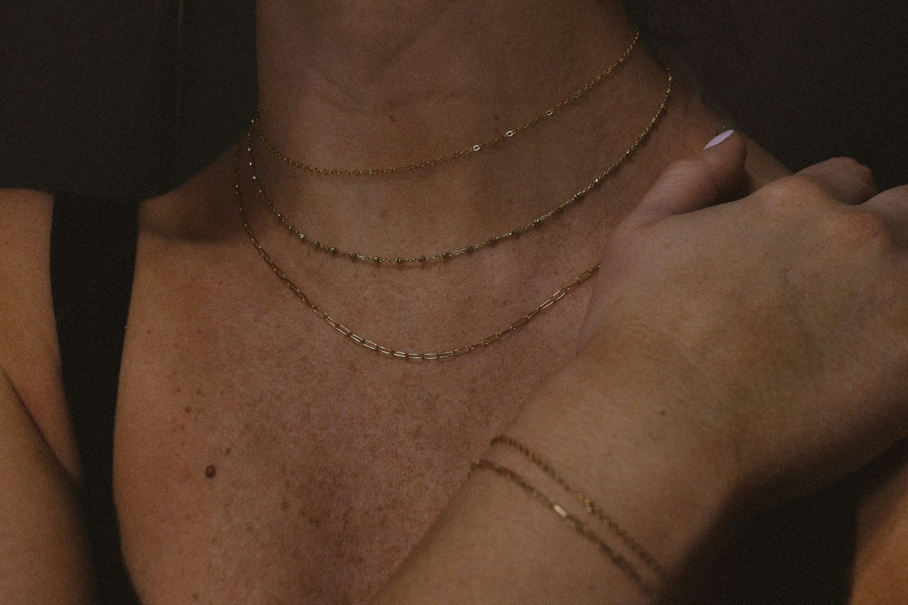 A close up of a yellow gold thin layered chain necklace on a woman’s neck and two bracelets of the same style on her wrist.