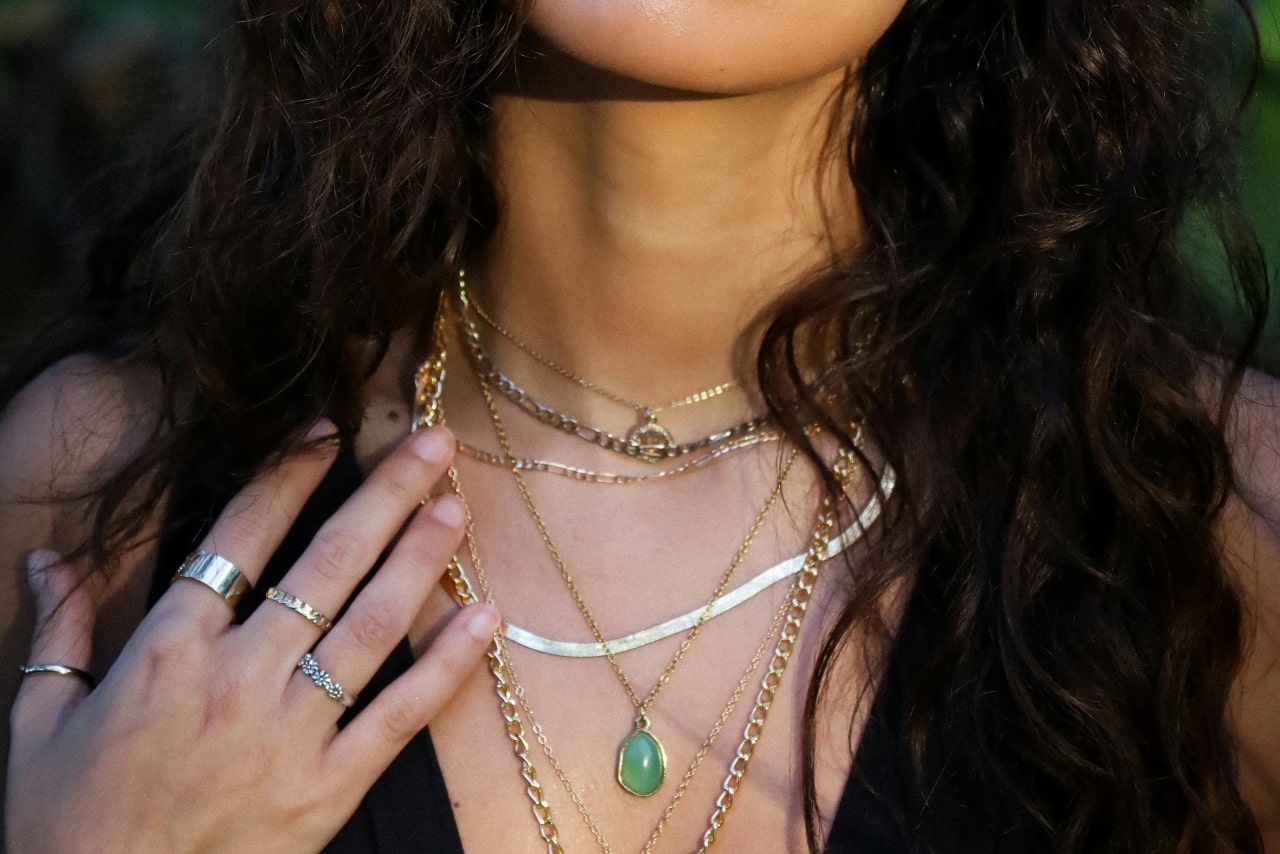A close up of layered necklaces and fashion rings on a woman with black curly hair.
