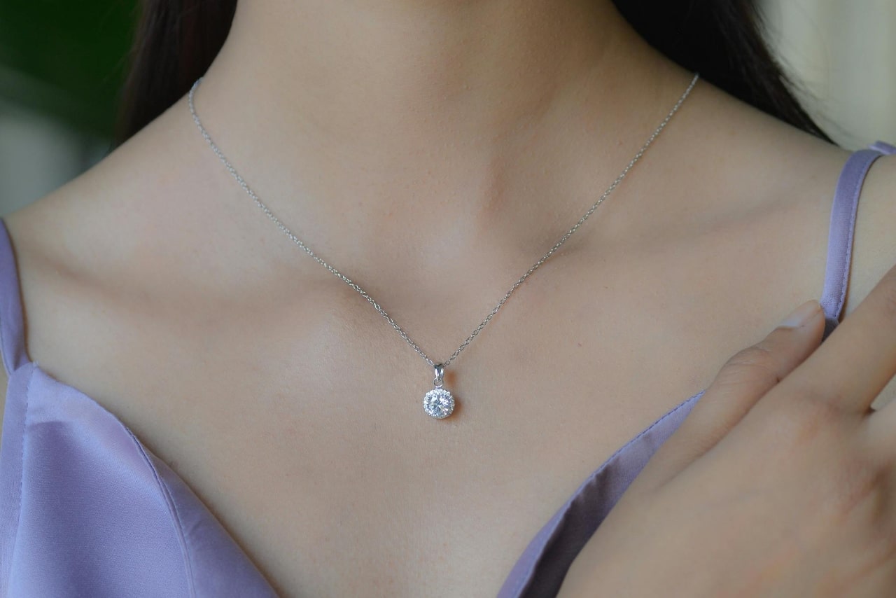 A close up of a white gold diamond pendant on a woman’s neck, dressed in a lavender satin top.