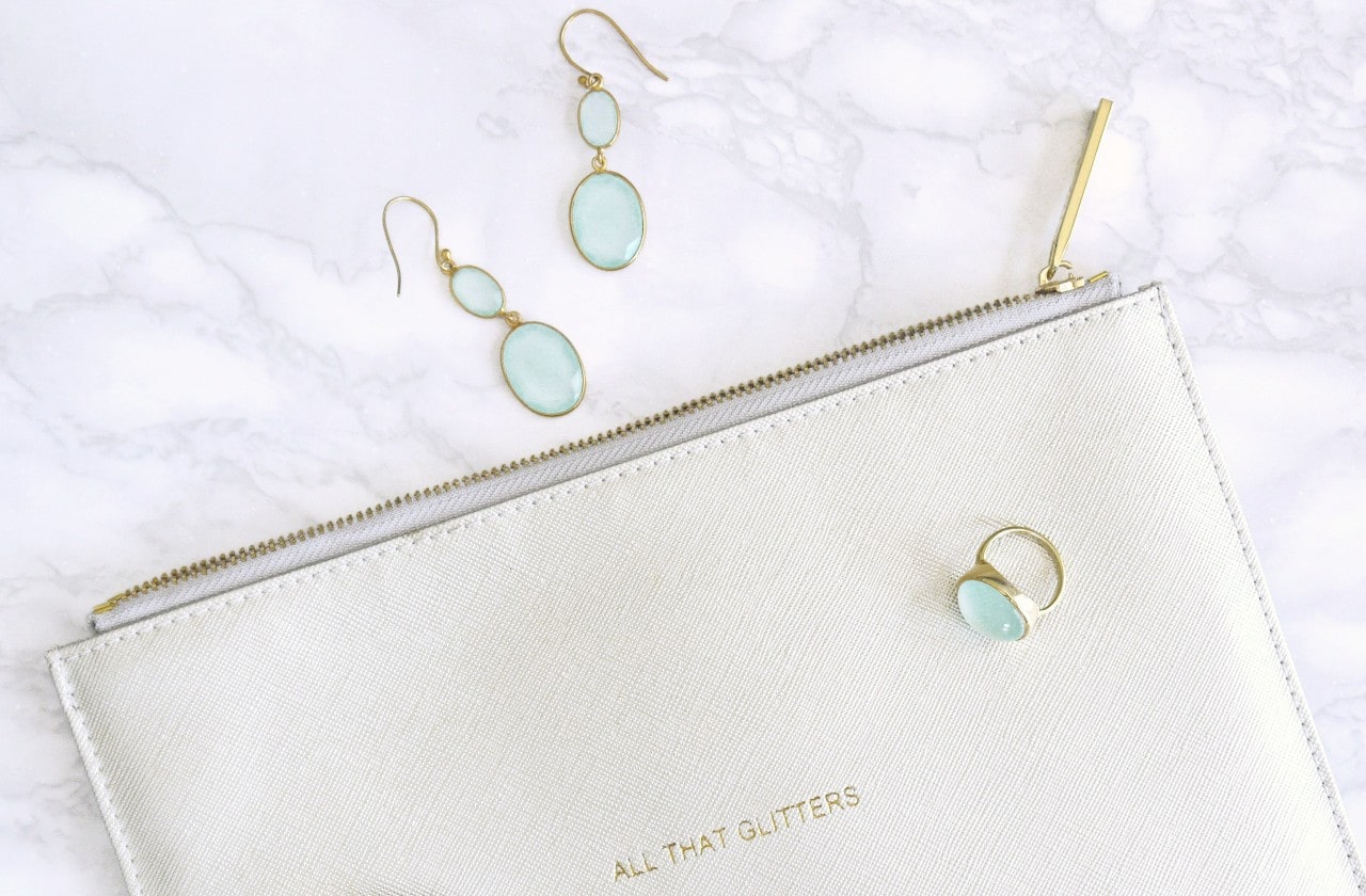 A pair of aquamarine and yellow gold earrings along with a matching fashion ring on top of a clutch on a white marble counter that says All That Glitters on it A pair of aquamarine and yellow gold earrings along with a matching fashion ring on top of a clutch on a white marble counter that says All That Glitters on it
