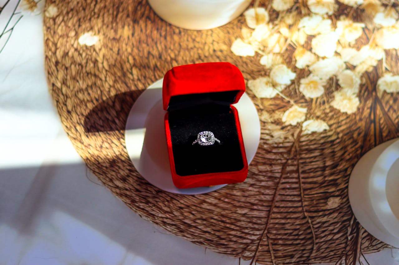 A close up of a halo white gold engagement ring inside the black and red velvet jewelry box placed on a textured surface.
