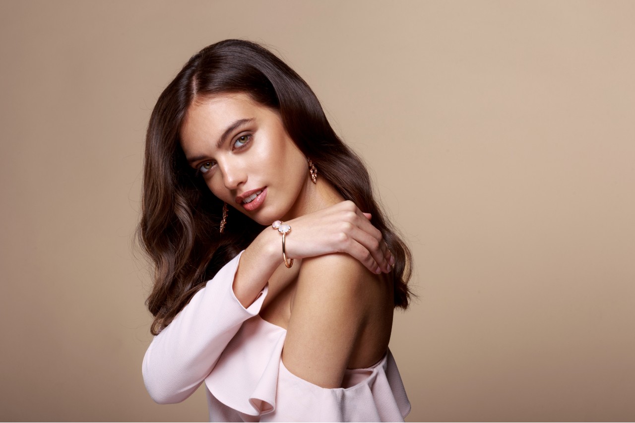 A woman with brown wavy hair poses in a blush off-shoulder top, showcasing a floral rose gold bracelet and matching earrings. A woman with brown wavy hair poses in a blush off-shoulder top, showcasing a floral rose gold bracelet and matching earrings.