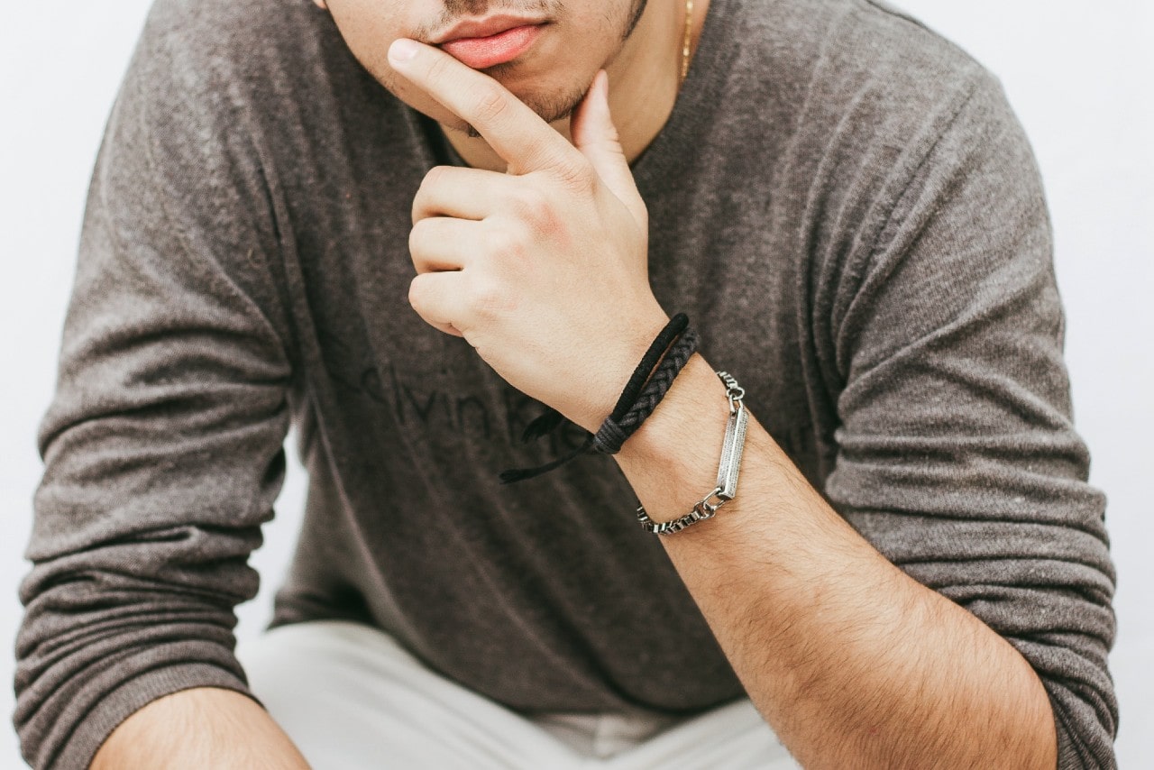 A man in a gray long sleeve sits, showcasing a variety of ribbon and metallic designer bracelets