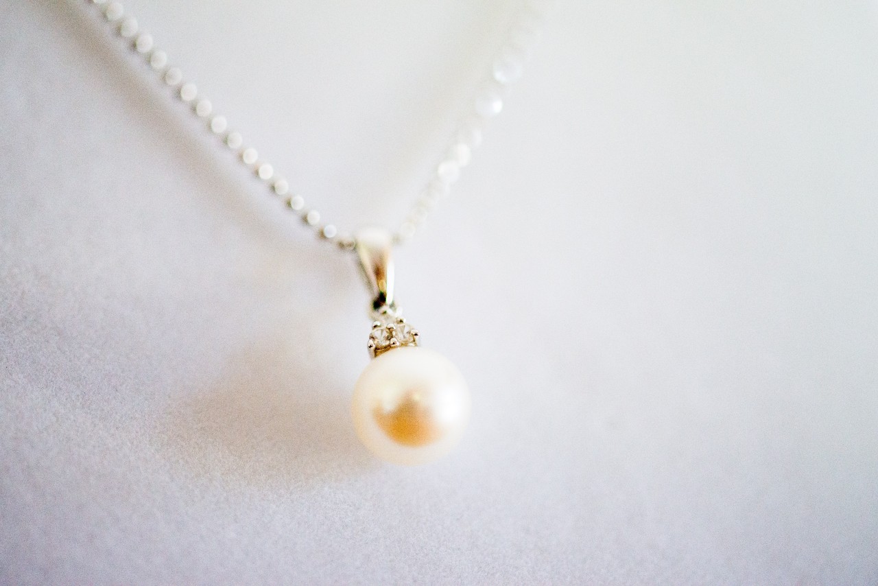 a pearl necklace on a white gold chain hangs by a white textile surface.
