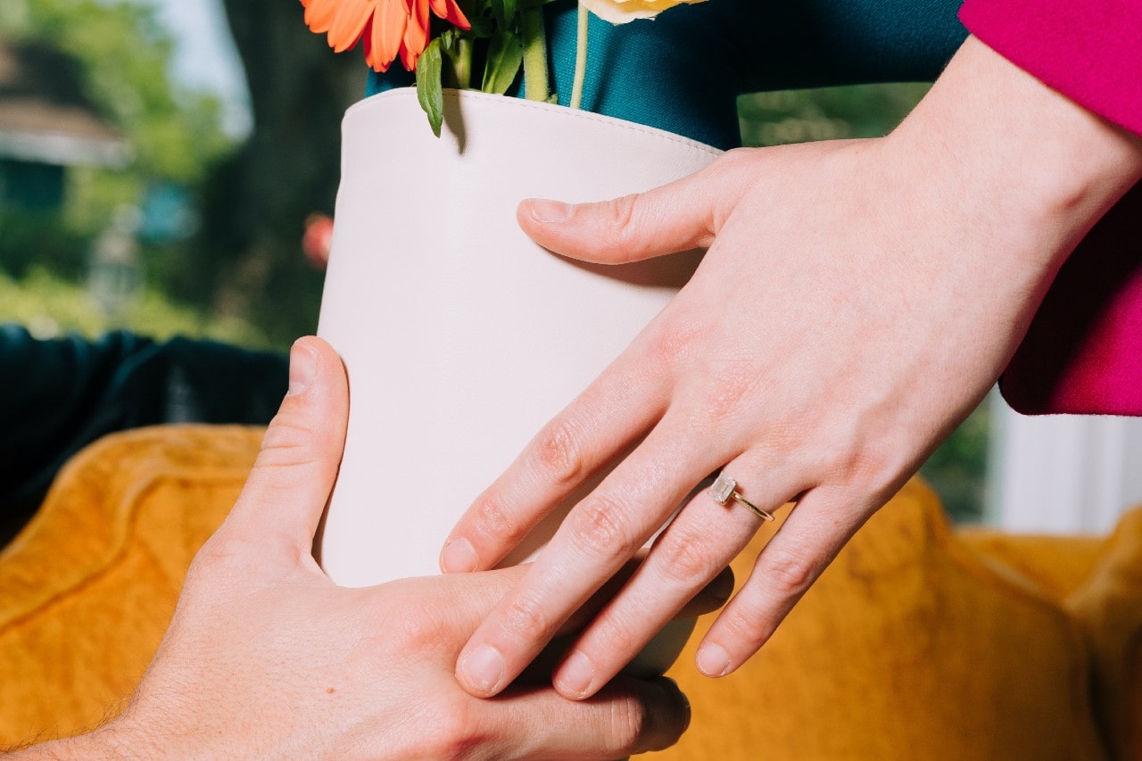 A man and woman passing a flower pot, the woman wearing a radiant cut engagement ring
