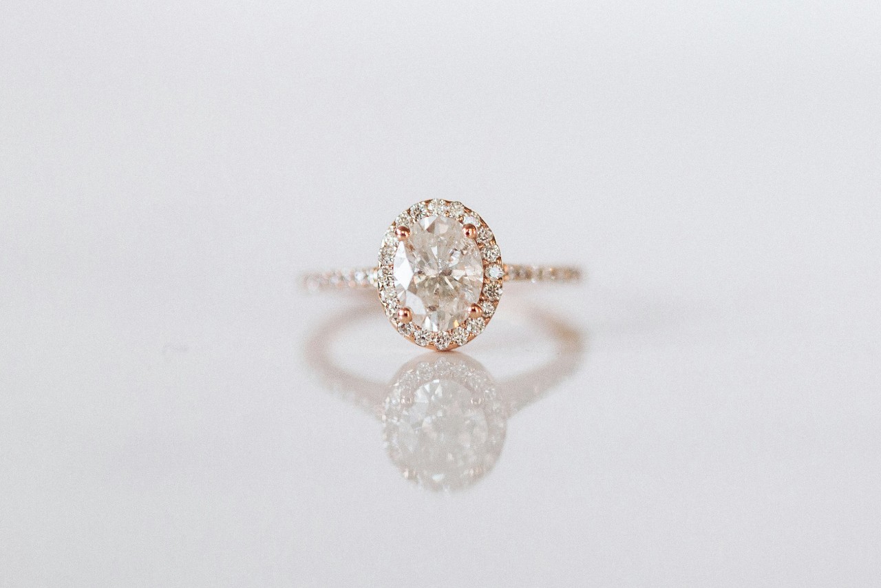A close up of a rose gold oval halo diamond engagement ring on the reflective surface