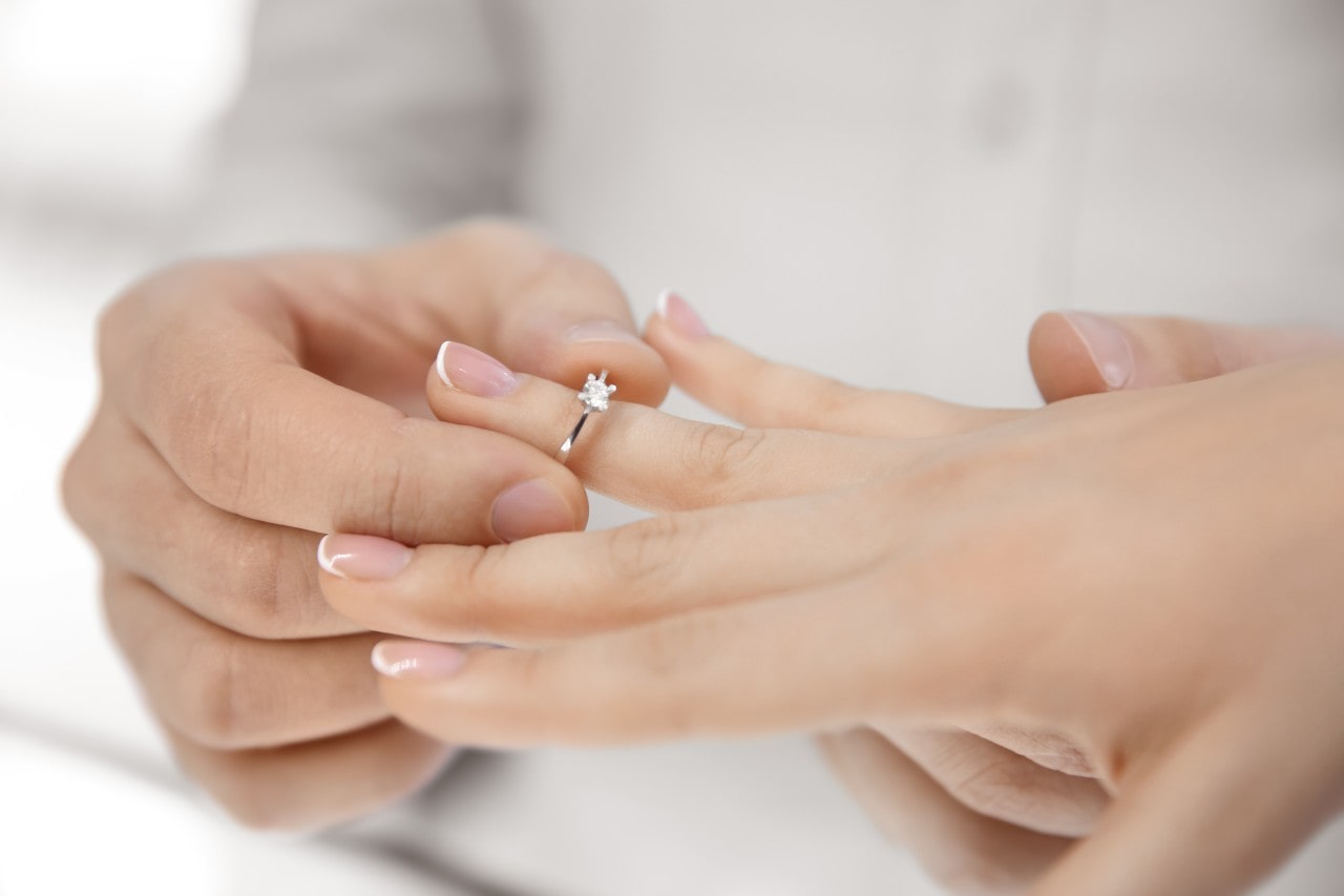 A close-up of a person’s hand putting on a yellow gold round cut diamond engagement ring on another person’s finger