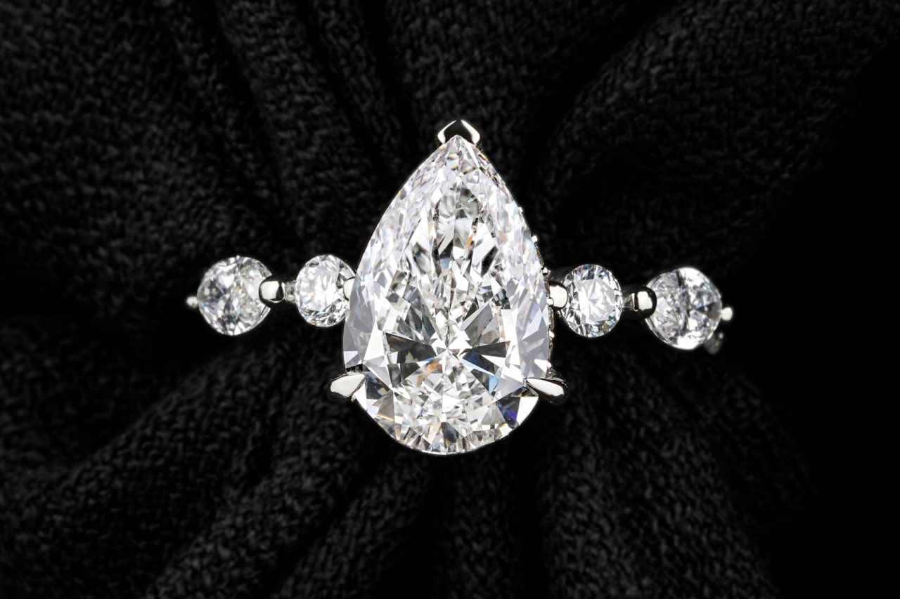 A close-up of an engagement ring with a pear shape center stone and round cut side stones. A close-up of an engagement ring with a pear shape center stone and round cut side stones.